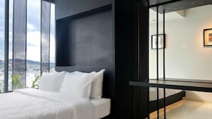 A modern bedroom with a bed, a black nightstand, and a large window offering a city view. - Pause Apartment (Dalat)