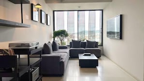 A modern living room with a large window, a sofa, a coffee table, and a shelving unit with books and decorative items. - Pause Apartment (Dalat)