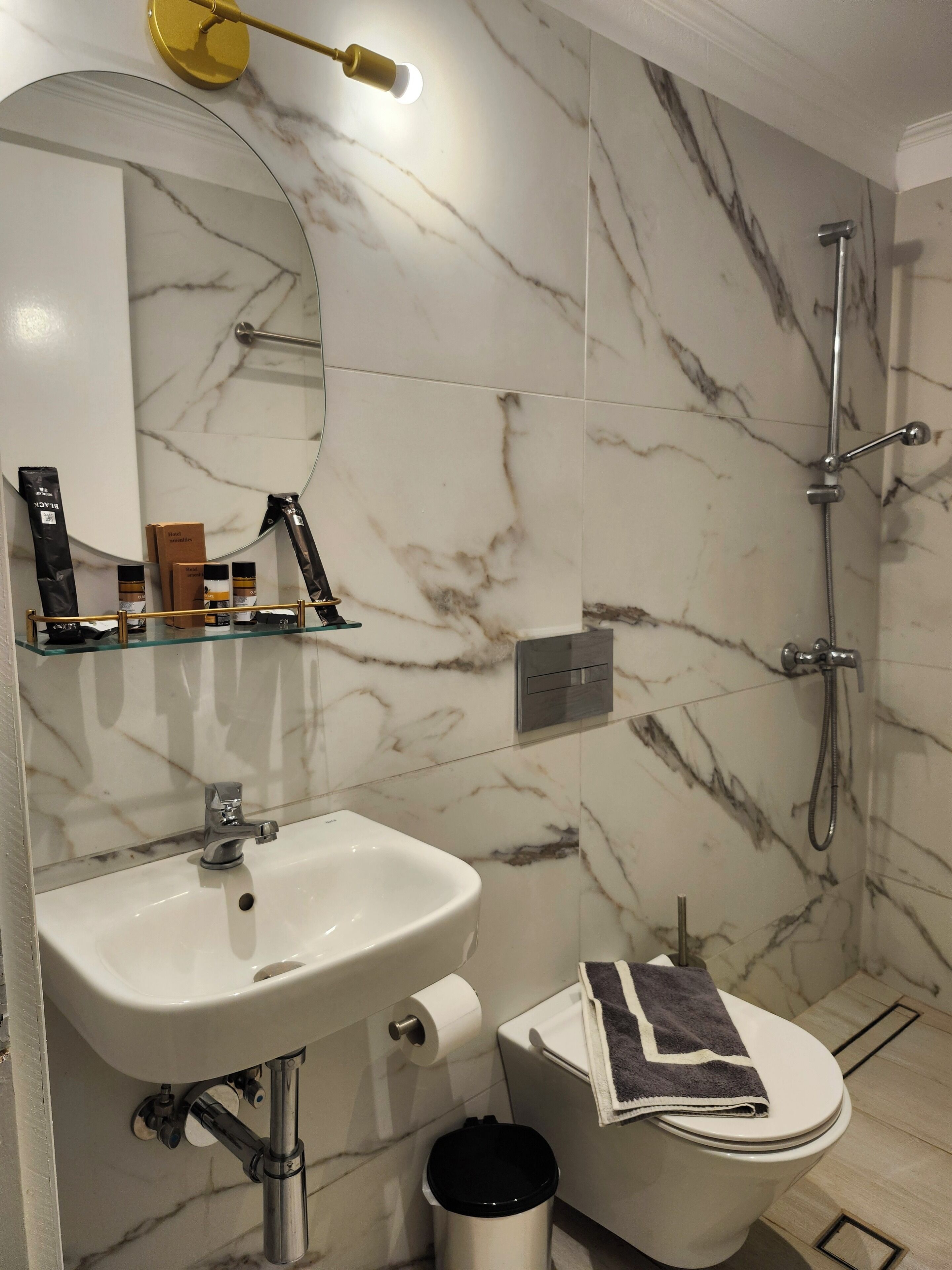 A bathroom with a white sink, a toilet, a shower, and a mirror with a gold light fixture.