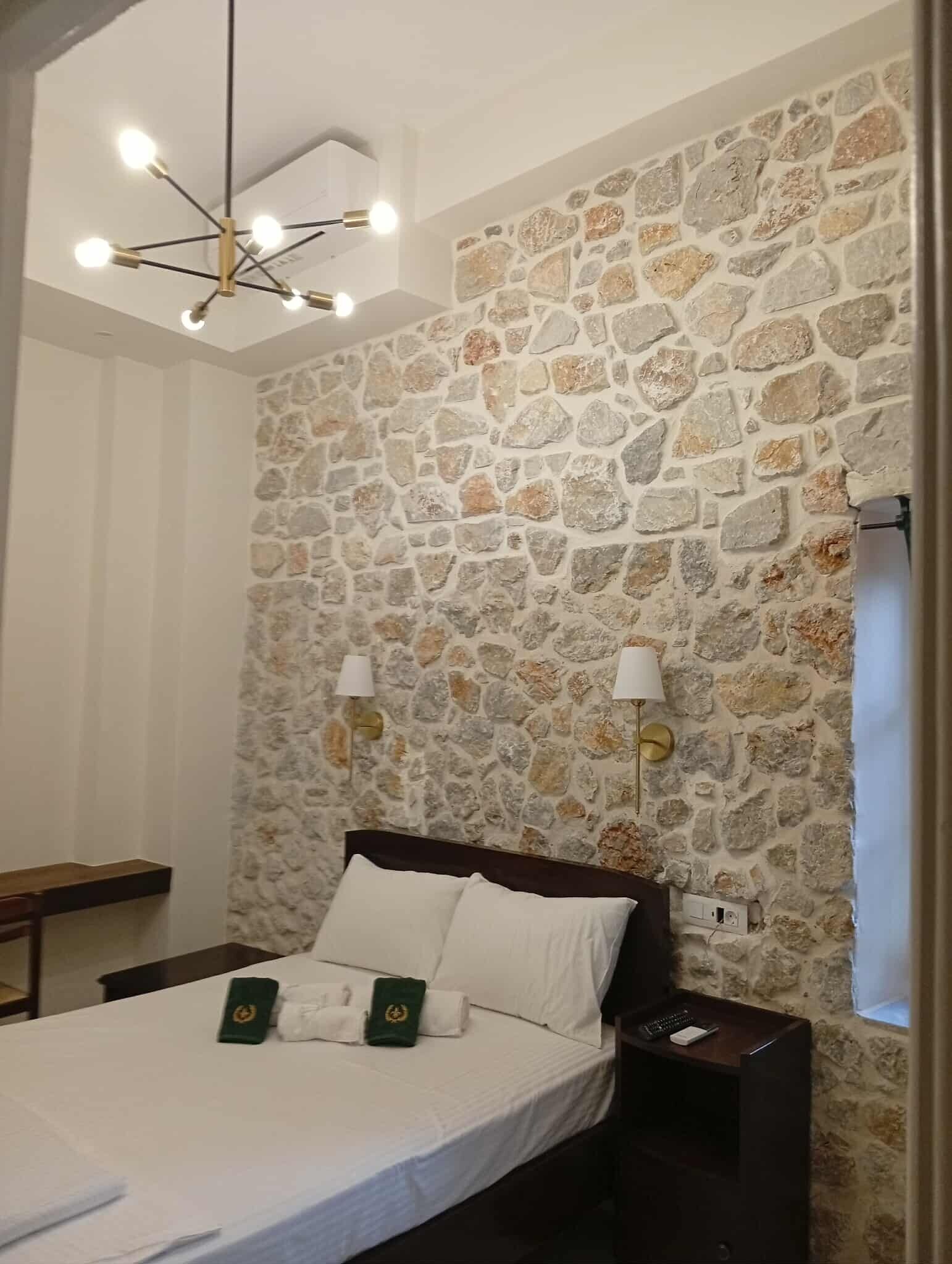 A bedroom with a stone wall, a bed with white linens, two bedside lamps, and a modern chandelier.
