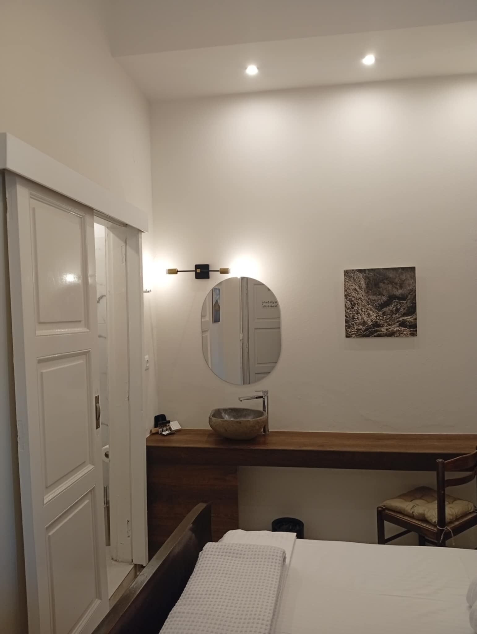A bedroom with a bed, a wooden dresser, a round mirror, a small sink, and a wall-mounted light fixture.
