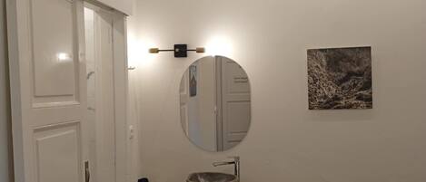 A bedroom with a bed, a wooden dresser, a round mirror, a small sink, and a wall-mounted light fixture.