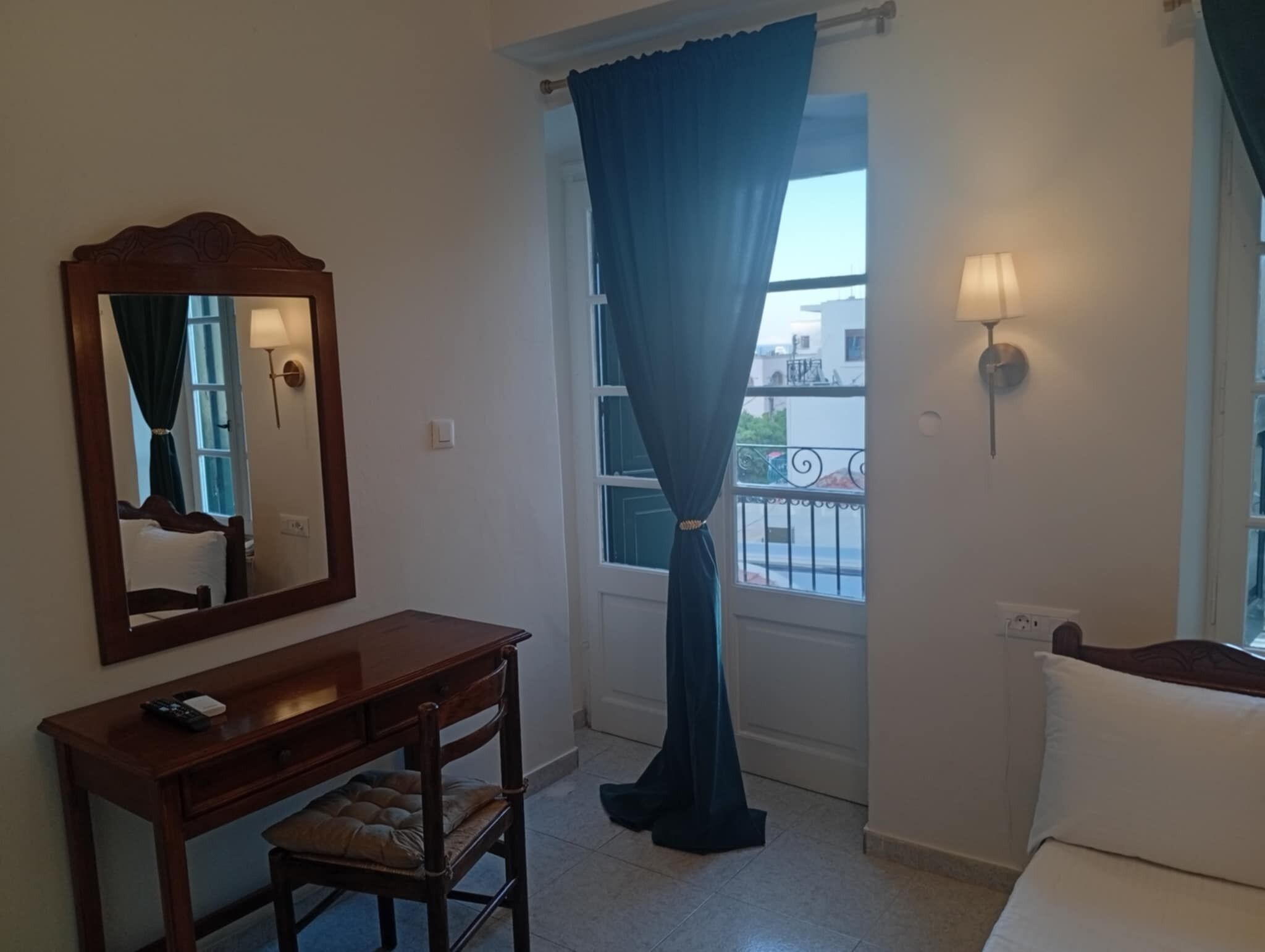 A room with a wooden desk, a chair, a bed, a window with curtains, and a wall-mounted lamp.