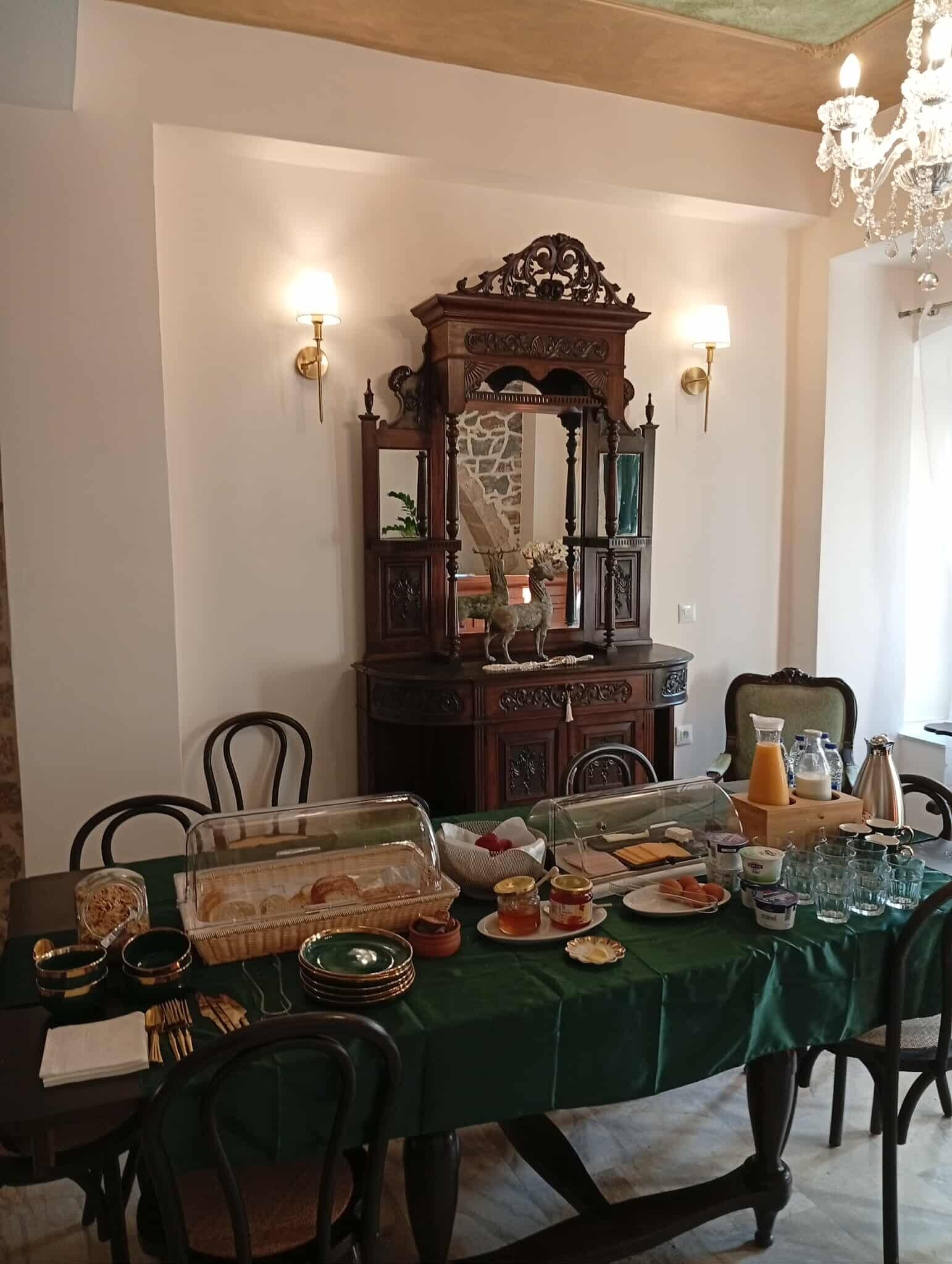 A dining area with a green tablecloth, wooden chairs, a decorative mirror, and various breakfast items.