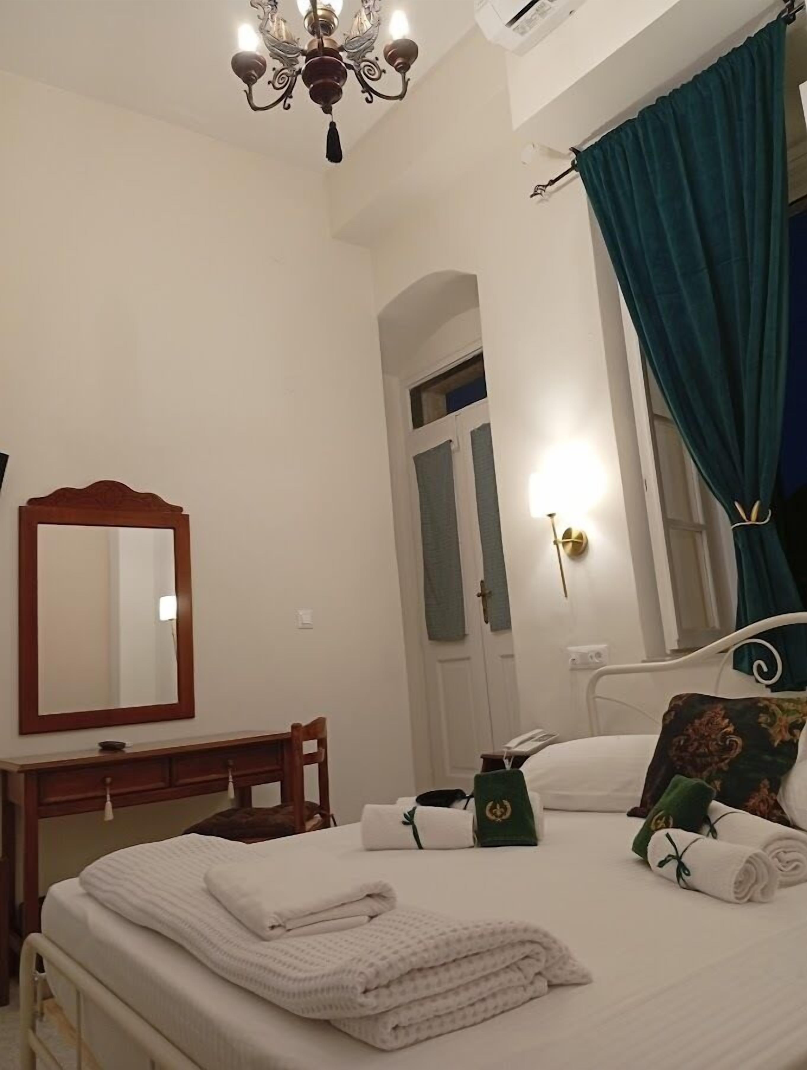 A hotel room with a bed, a bedside table, a mirror, a chandelier, a door, and a window with curtains.