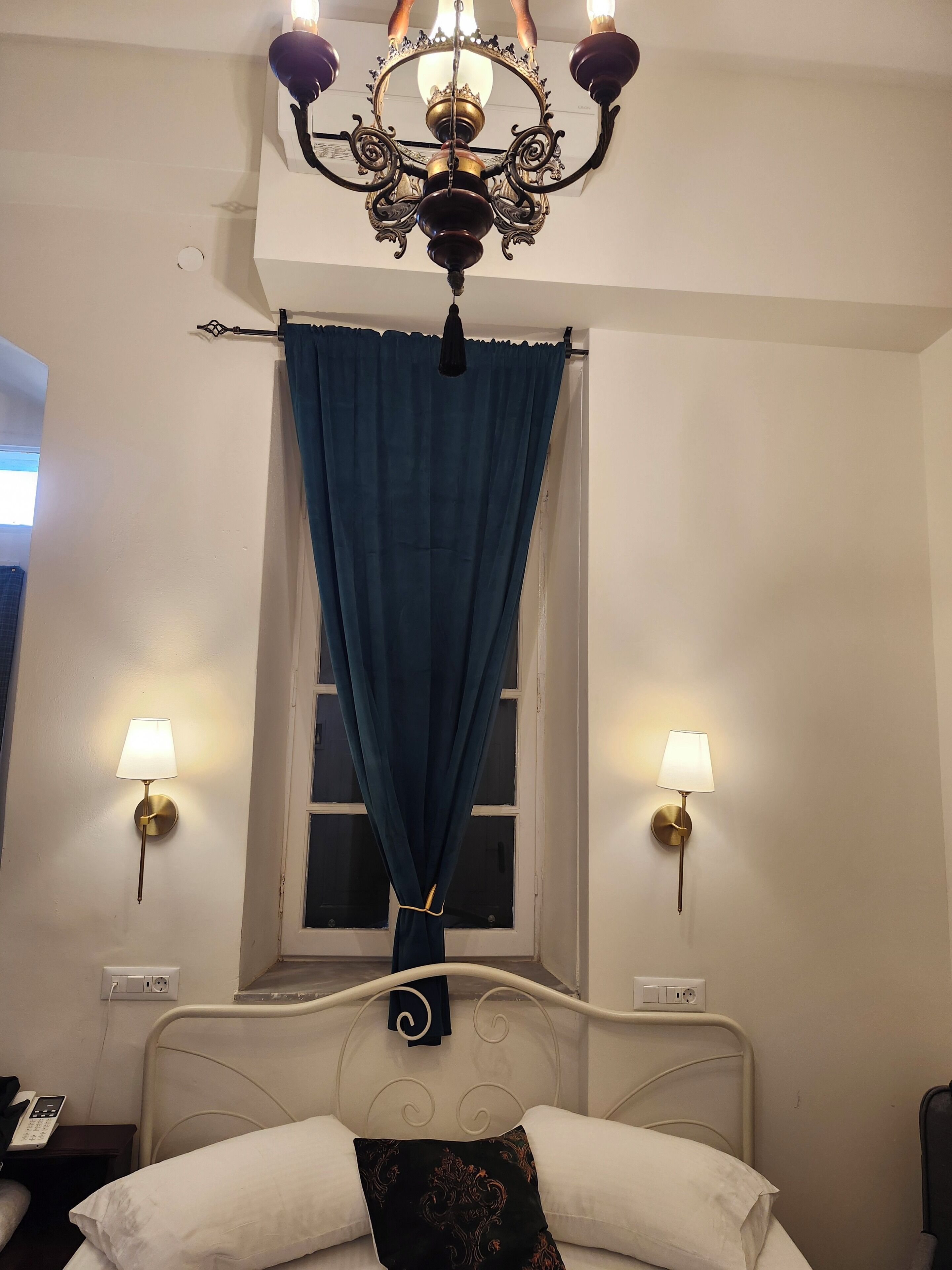 Une chambre d’hôtel avec un lit, un lustre, deux lampes fixées au mur et une fenêtre avec des rideaux.