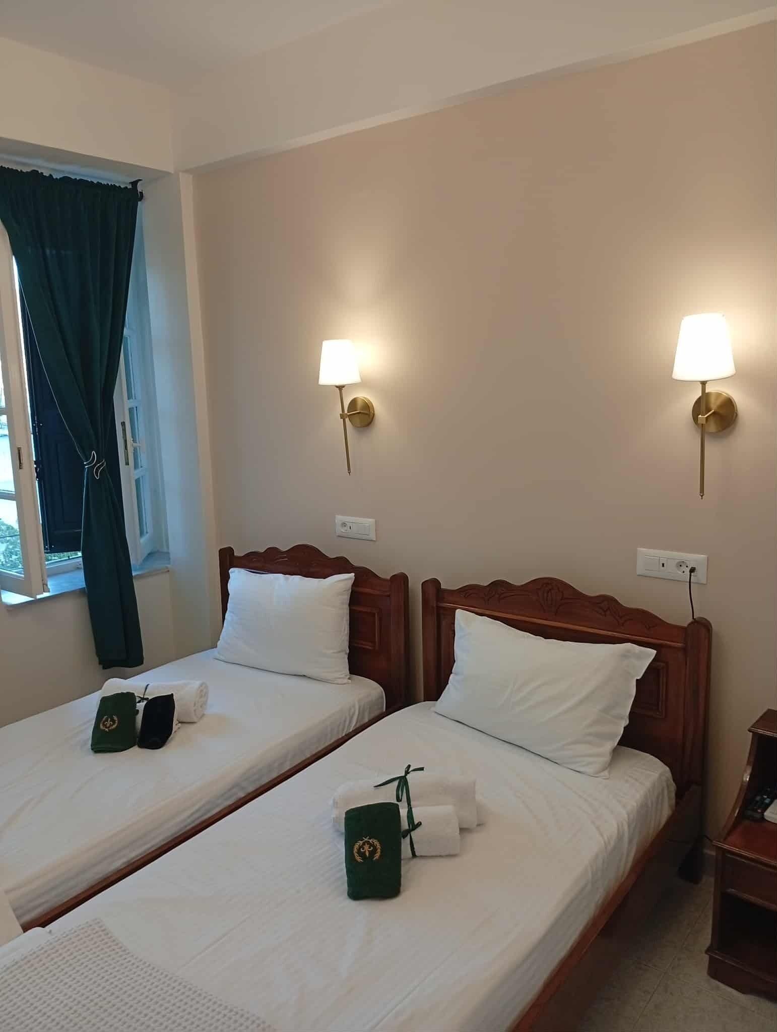 Habitación de hotel con dos camas individuales, cada una con cabecero de madera, ropa blanca y un bolso verde sobre una cama. Hay dos lámparas de pared con detalles dorados.