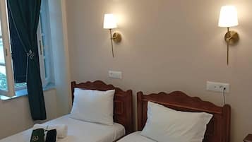 A hotel room with two single beds, each with a wooden headboard, white linens, and a green bag on one bed. There are two wall-mounted lamps with gold fixtures.