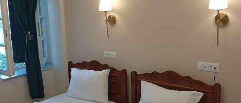 Habitación de hotel con dos camas individuales, cada una con cabecero de madera, ropa blanca y un bolso verde sobre una cama. Hay dos lámparas de pared con detalles dorados.