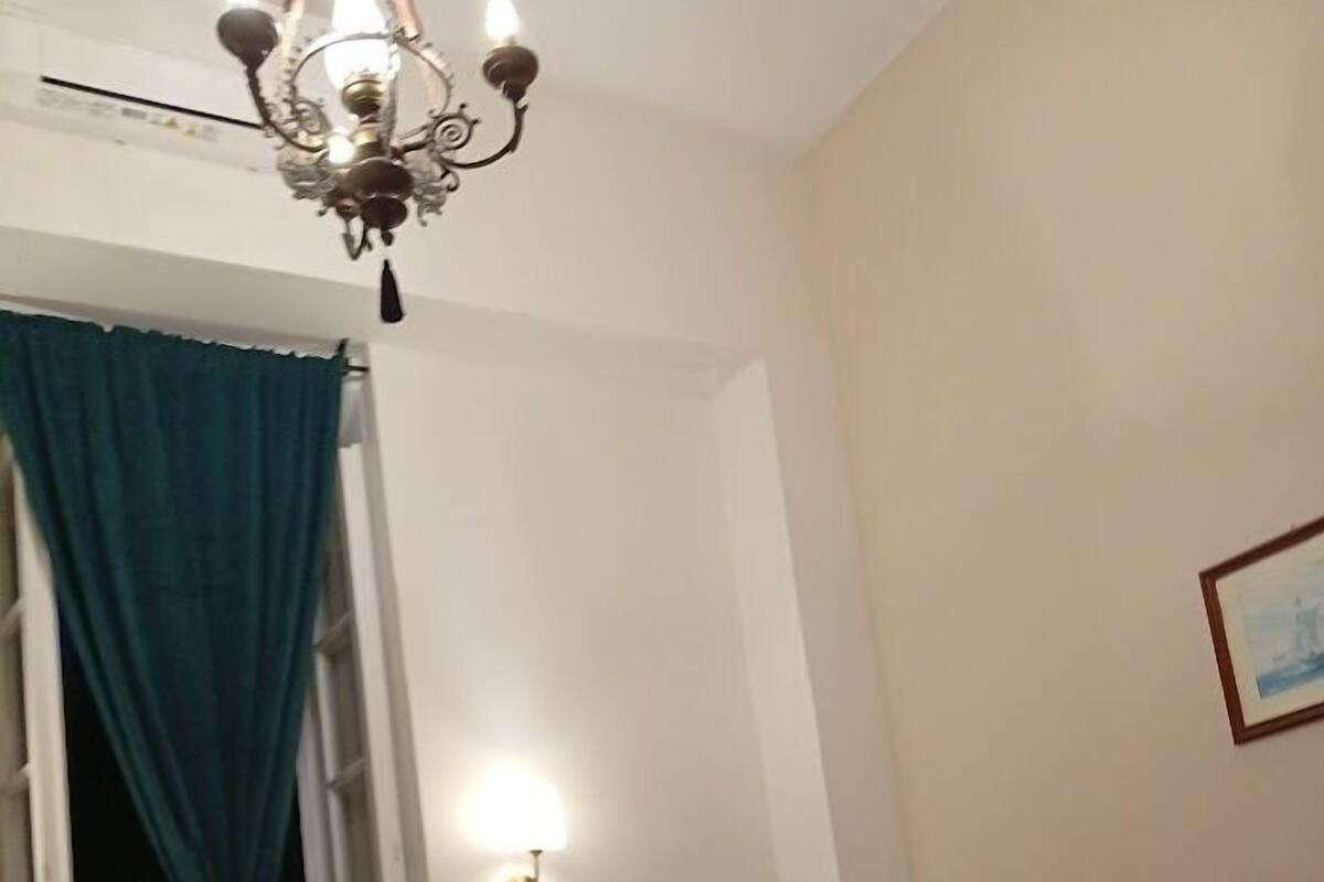 Habitación de hotel con cama, una silla, un candelabro, una ventana con cortinas y una lámpara de pared.