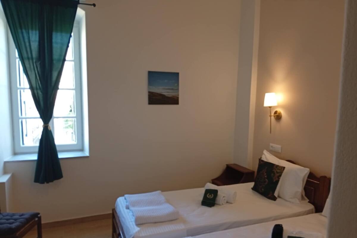 Habitación de hotel con dos camas, un escritorio, una silla, una ventana con cortinas, una lámpara de pared y un cuadro pequeño.