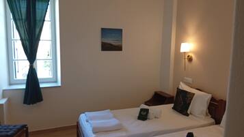 A hotel room with two beds, a desk, a chair, a window with curtains, a wall-mounted lamp, and a small painting.