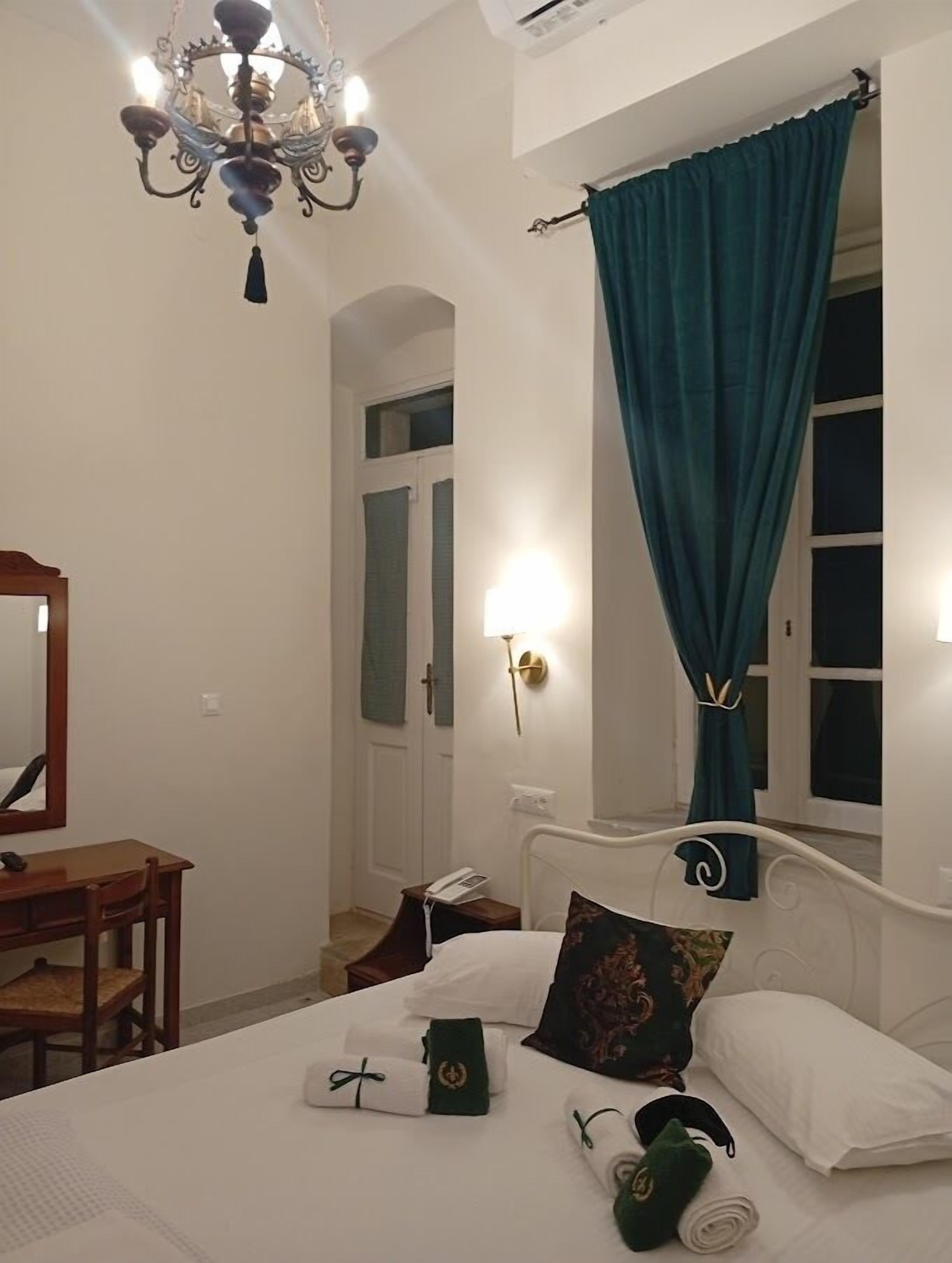 Quarto de hotel com cama, escrivaninha, cadeira, lustre, janela com cortinas e luminária na parede.