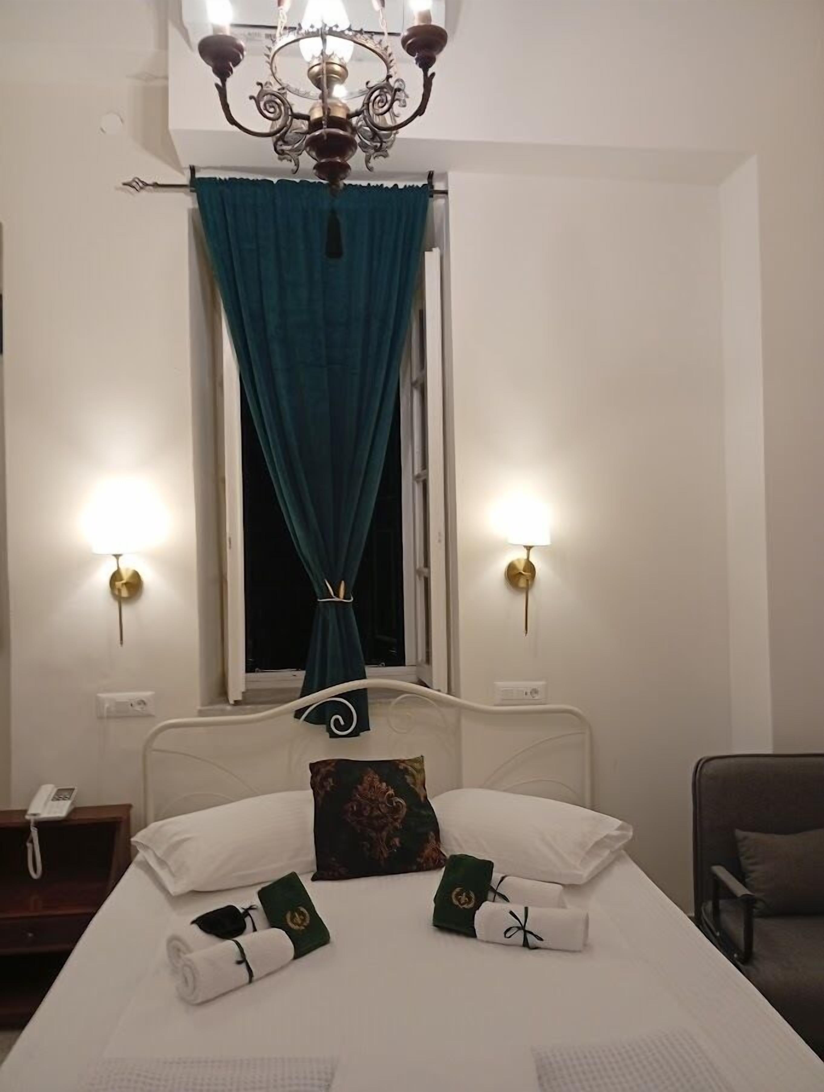 A hotel room with a bed, a chandelier, a window with curtains, bedside lamps, and a chair.