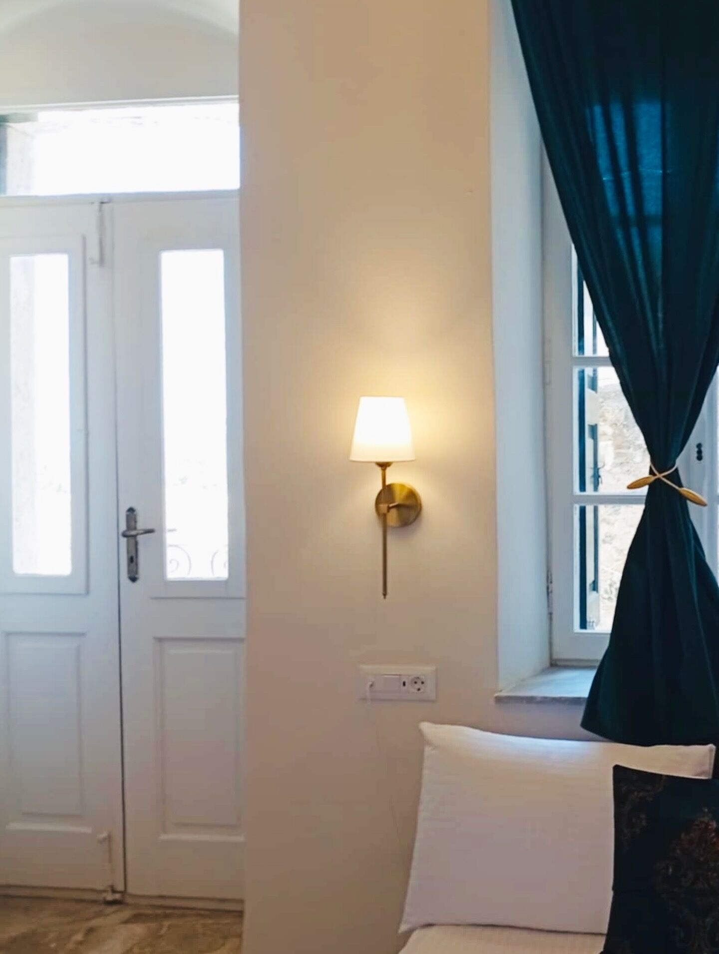 A room with a bed, a wall-mounted lamp, a window with dark curtains, and a door.