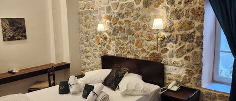 Habitación de hotel con pared de piedra, cama con ropa de cama blanca, mesita de noche con lámpara, teléfono y ventana con cortinas.