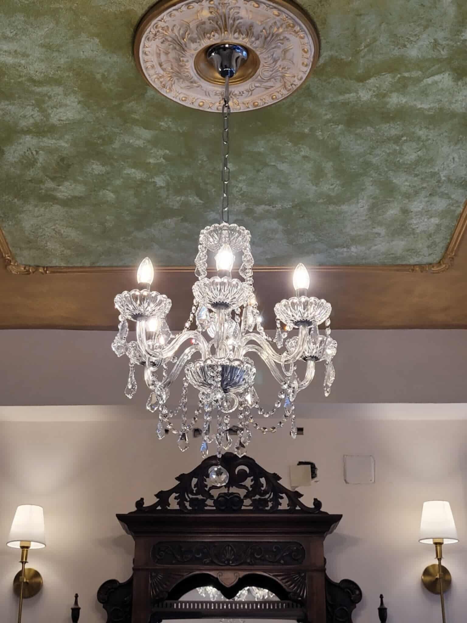 Un candelabro con múltiples brazos y luces de cristal, un medallón decorativo en el techo y dos lámparas de pared.