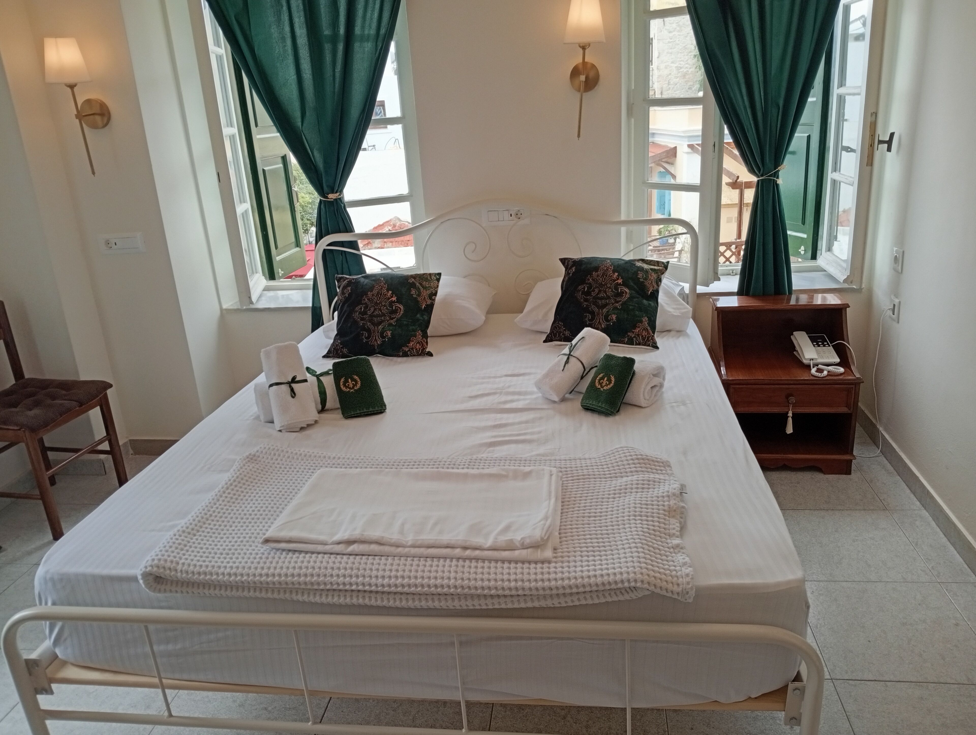 A neatly made bed with towels and pillows, a bedside table with a phone, and a chair in a room with windows and curtains.