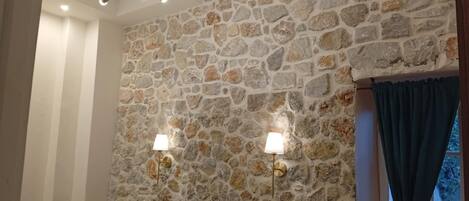 Un dormitorio con pared de piedra, cama con ropa de cama blanca, dos lámparas de pared y una lámpara en el techo.