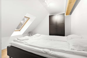 Ein Schlafzimmer mit schrägen Wänden, einem Oberlicht, einem Bett mit weißer Bettwäsche, einem dunklen Kleiderschrank und einem kleinen runden Stuhl.