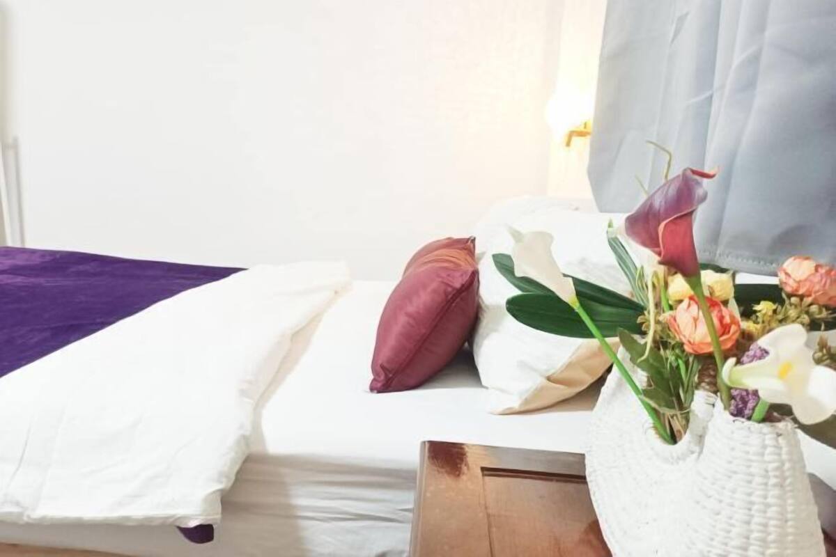 A bed with white and purple bedding, a wooden headboard, and a vase with flowers.