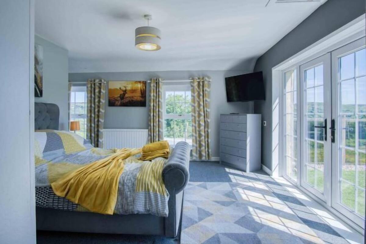 Un dormitorio con cama, cómoda, televisor y un amplio ventanal con cortinas.