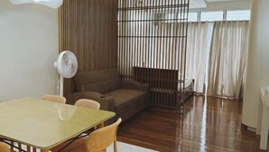 A living room with a glass table, wicker chairs, a brown sofa, a white fan, and a wooden partition. - Queensland Manor Condominium Deluxe Suite (Cebu City)