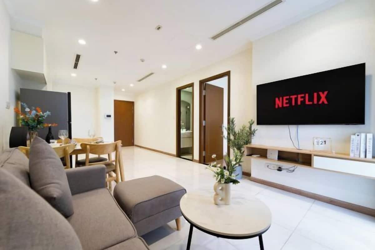 A modern living room with a sofa, a coffee table, a TV displaying 'NETFLIX,' and a dining area with a table and chairs.