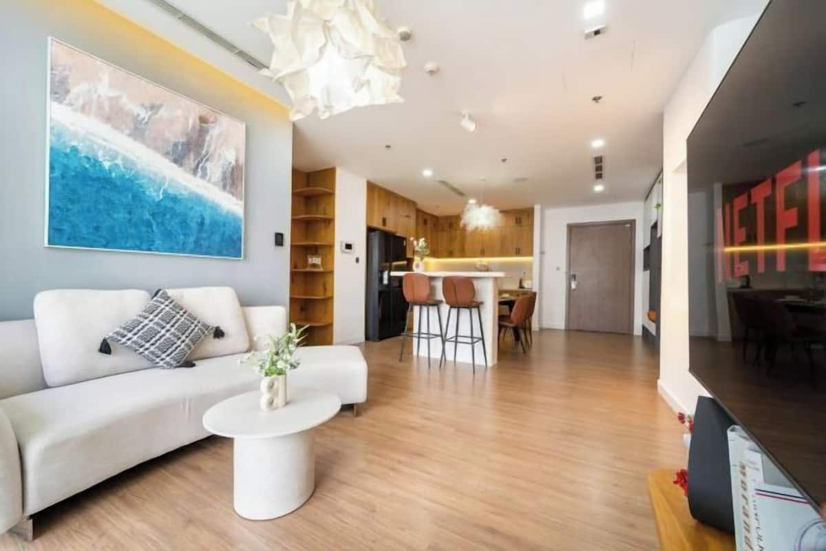 A modern living room with a sofa, coffee table, and a painting. There is a kitchen area with wooden cabinets and a dining space with chairs.