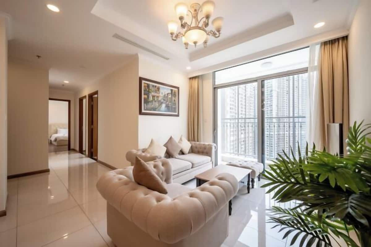 A spacious living room with a large window, a chandelier, and a sofa with cushions.