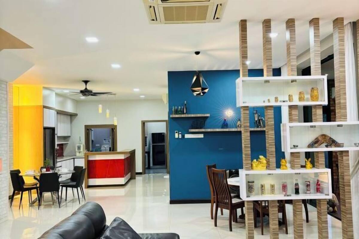 A modern living room with a sofa, a dining area with chairs, a kitchen in the background, and a decorative shelf with items.