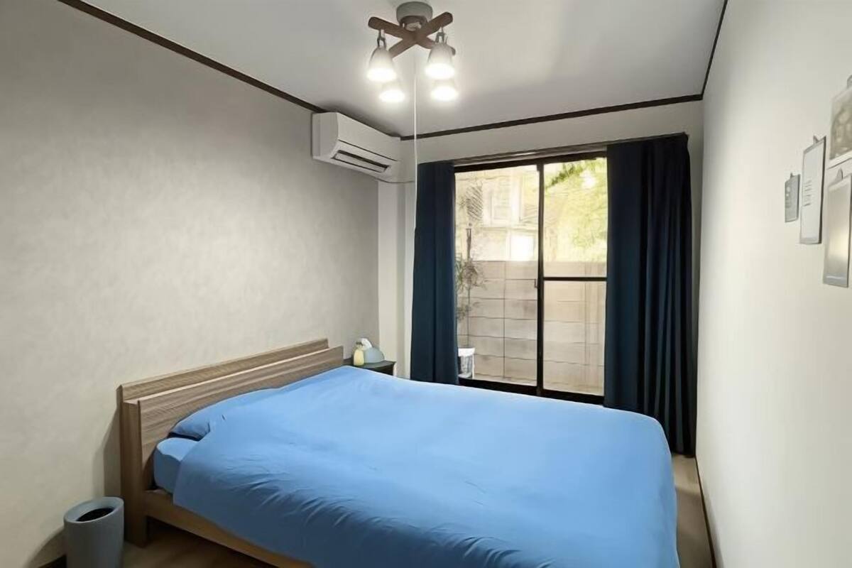 Una cama individual con colchón azul en una habitación con ventana, unidad de aire acondicionado y ventilador de techo.