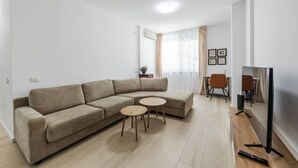 Una sala de estar moderna con un sofá seccional beige, dos mesitas de madera, un televisor de pantalla plana y un comedor con una mesa y sillas.