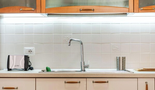 A kitchen with white tiles, a stainless steel faucet, a toaster, and wooden cabinets. - Sophie Downtown Apartment Modern 1BR (Tirana)