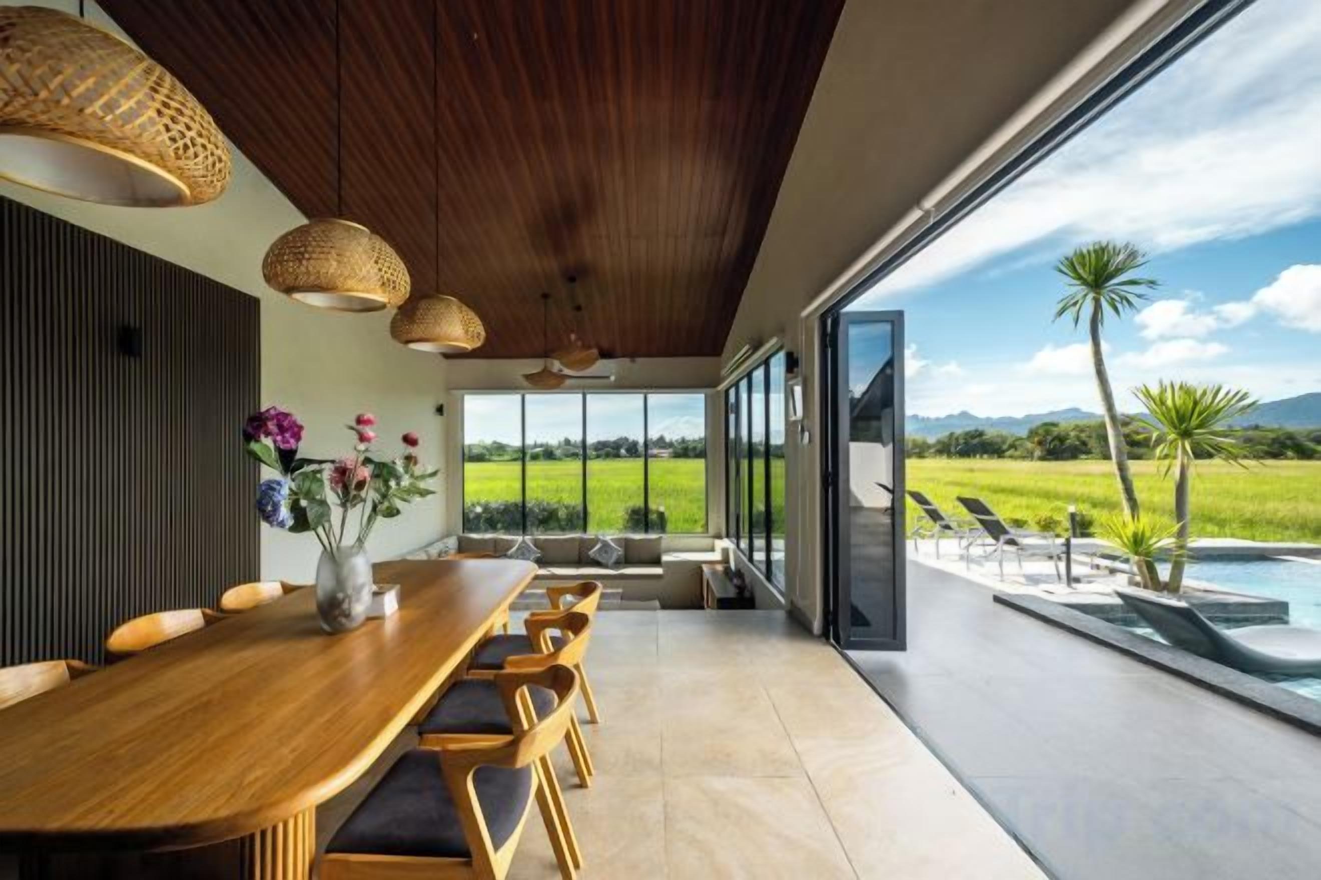 A modern dining area with a long wooden table, wicker pendant lights, and a view of a swimming pool and landscape.