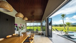 A modern dining area with a wooden table and chairs, large windows offering a view of a pool and landscape. - Villa Moo Moo (Langkawi)