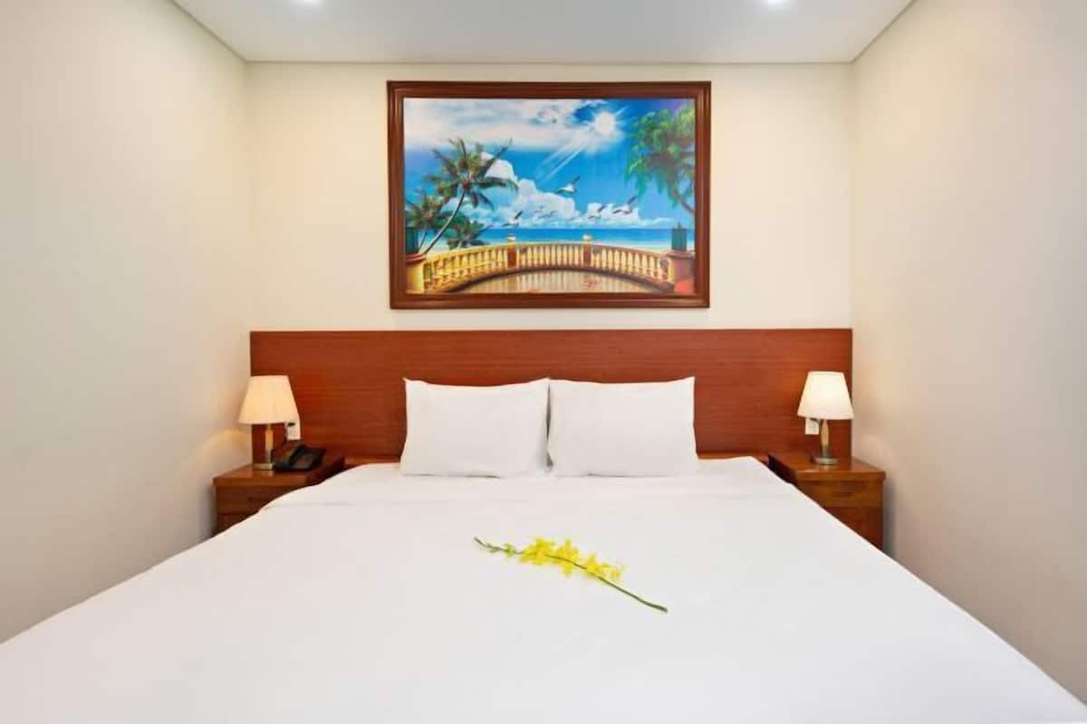 Una camera d'albergo con un letto, due comodini con lampade, un quadro raffigurante una spiaggia e un singolo fiore giallo sul letto.