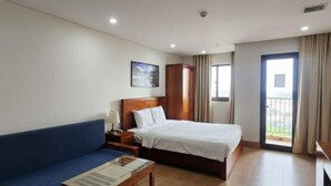 A hotel room with a bed, a blue sofa, a wooden nightstand, a table with a lamp, a painting on the wall, and a balcony door. - Cherry Apartment & Hotel (Da Nang)