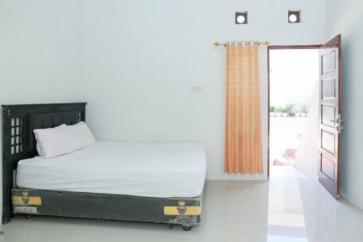 A single bed with a dark headboard and white bedding in a room with a white wall and a door leading outside.