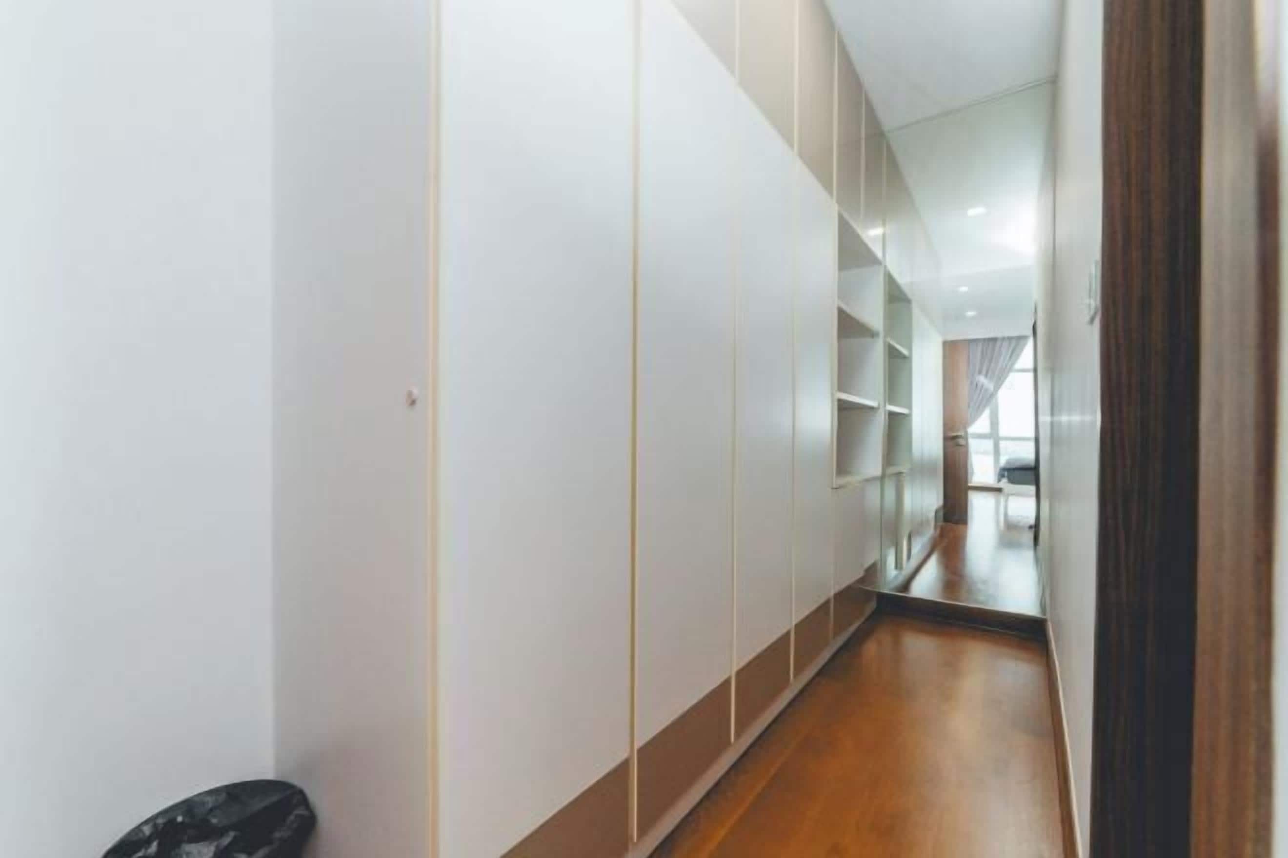 A narrow hallway with white walls and wooden flooring, leading to a room with a large window.