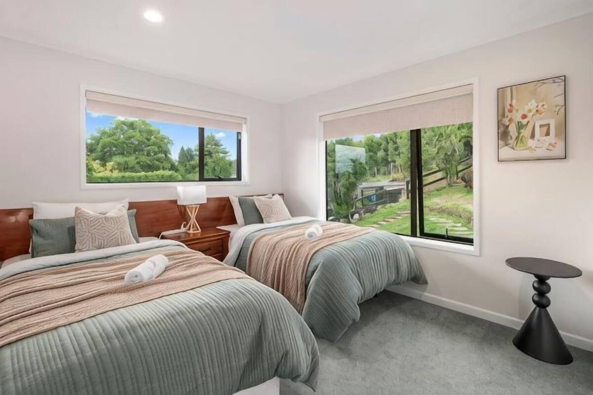 A bedroom with two beds, a bedside table, a lamp, a round table, and large windows with a view of greenery.
