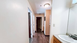 Un couloir étroit avec un plancher en bois, un mur blanc et une salle de bain équipée d’un lavabo et d’un miroir.