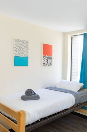 A single bed with a wooden frame, a white mattress, and a grey blanket. There are two wall-mounted artworks, one with a blue and white pattern and the other with a red and black pattern. A window with blue curtains is visible. - Private 2BR/2BA Downtown Apartment | River and Mou (Pittsburgh)