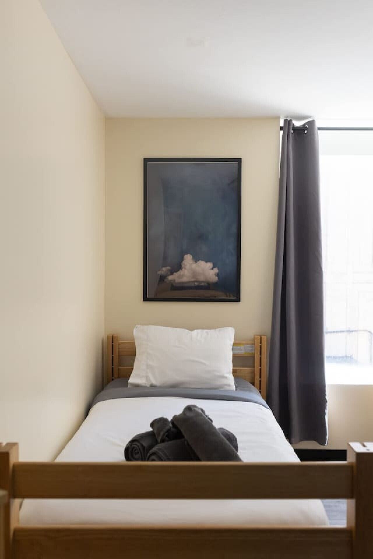 A single bed with a wooden frame, a white pillow, a rolled-up towel, and a dark curtain.