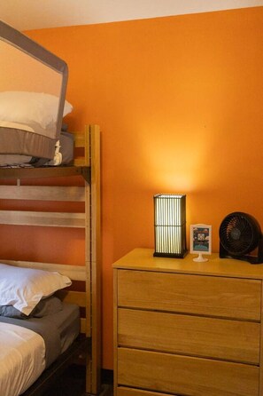 Un lit superposé avec une échelle, une table de chevet avec une lampe, un ventilateur et un petit tableau encadré.
