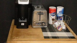 En kaffebryggare, en brödrost och en korg med burkar med kaffegrädde, placerade på ett träbricka mot en mörk vägg, med ett abstrakt konstverk i bakgrunden.