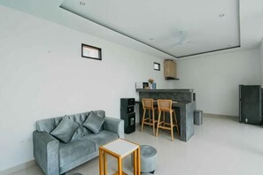 Ein modernes Wohnzimmer mit grauem Sofa, einem kleinen Holztisch und einer Küchenzeile mit einer Theke und Stühlen.