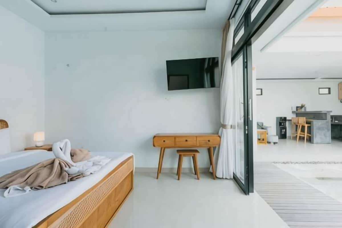 A modern hotel room with a bed, a desk, a television, and a view of the adjacent kitchen area.