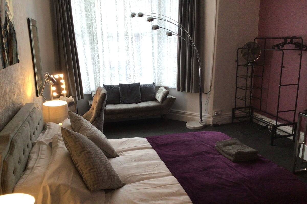 A neatly arranged bedroom with a bed, a sofa, a standing lamp, and a vanity with lights.