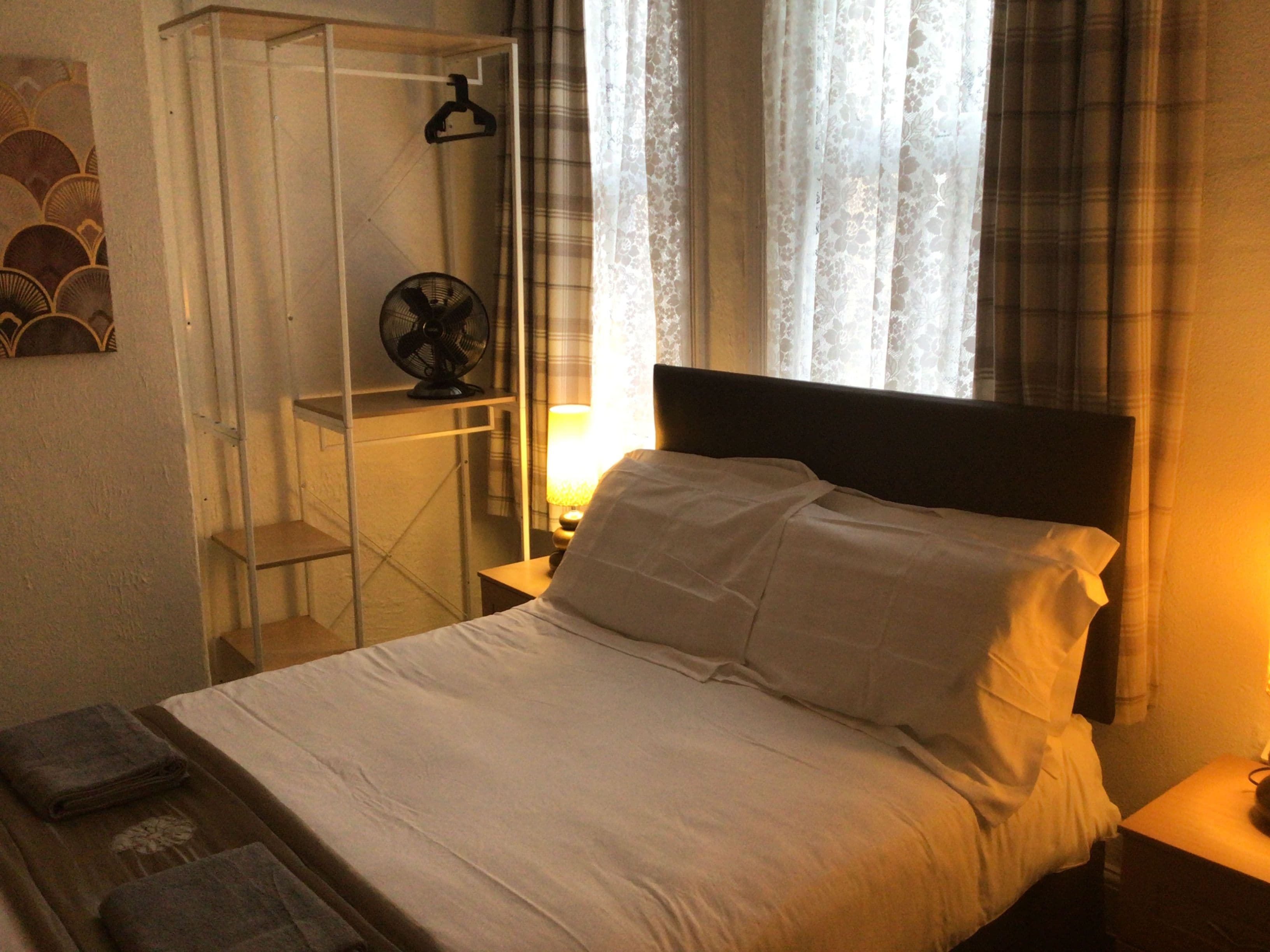 A neatly made bed with white linens, a bedside table with a lamp, a fan on a shelf, and a window with curtains.
